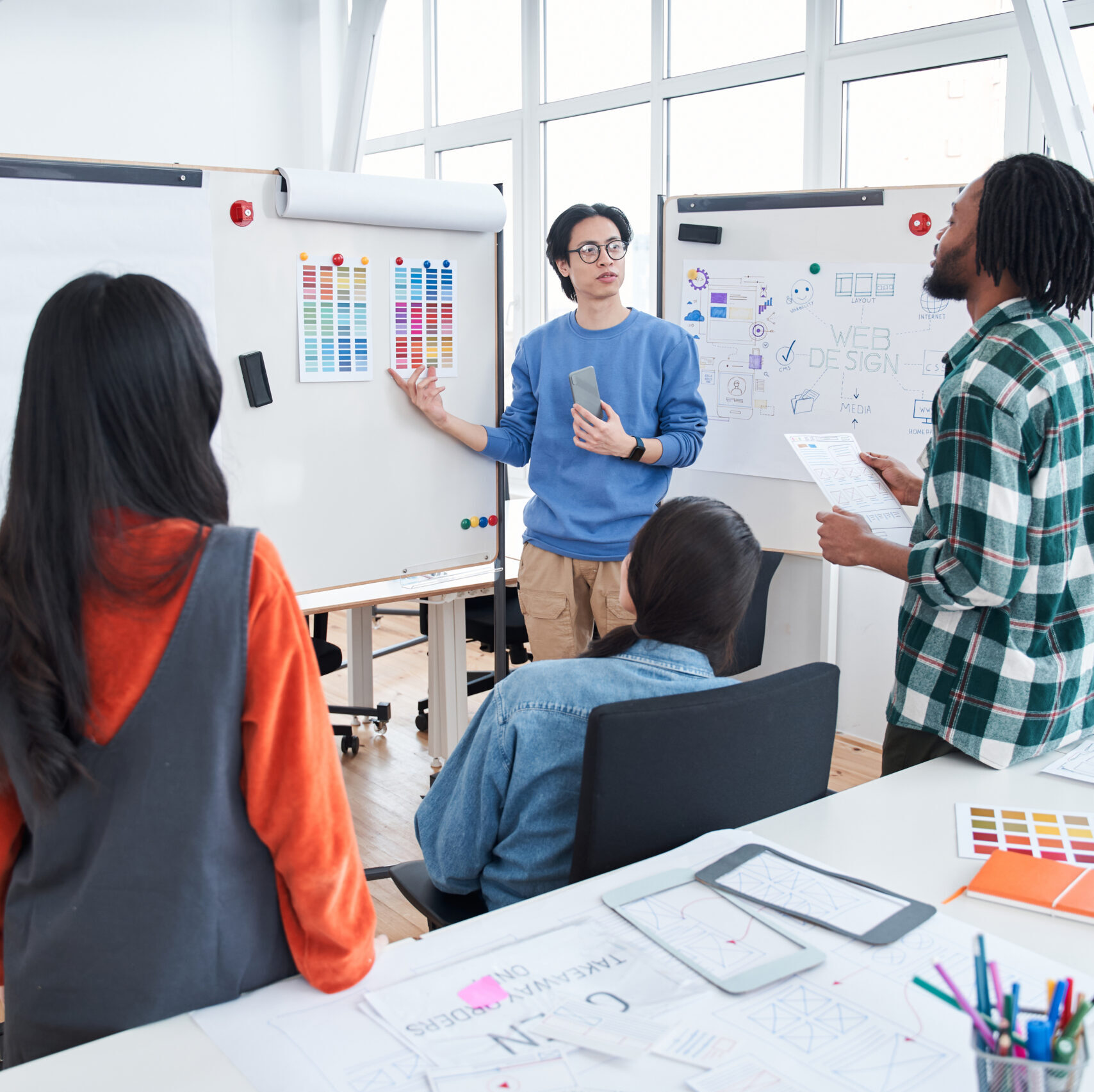 Students standing at the classroom and discussing project on interactive whiteboard. Handsome multiracial man wearing glasses pointing at the different colors while consulting with his classmates