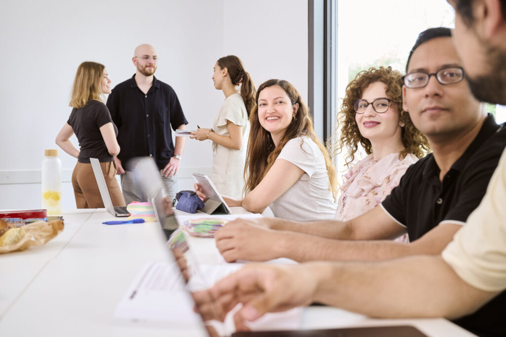 Eine Gruppe sich besprechender und arbeitender Personen, wovon vier an einem Tisch sitzen und die anderen drei im hinteren Teil des Raumes stehend ein Gespräch führen.
