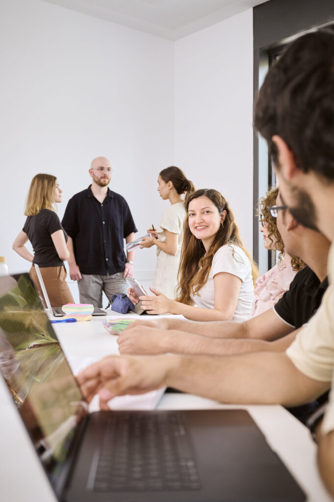 Eine Gruppe sich besprechender und arbeitender Personen, wovon vier an einem Tisch sitzen und die anderen drei im hinteren Teil des Raumes stehend ein Gespräch führen.