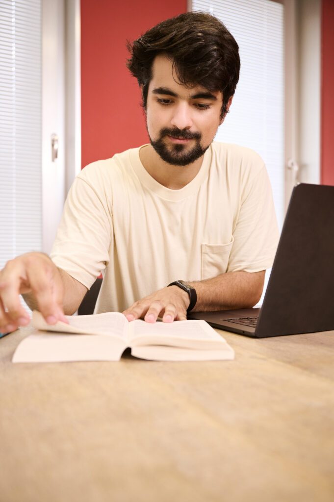 Student liest in Buch, sitzt vor dem Laptop.