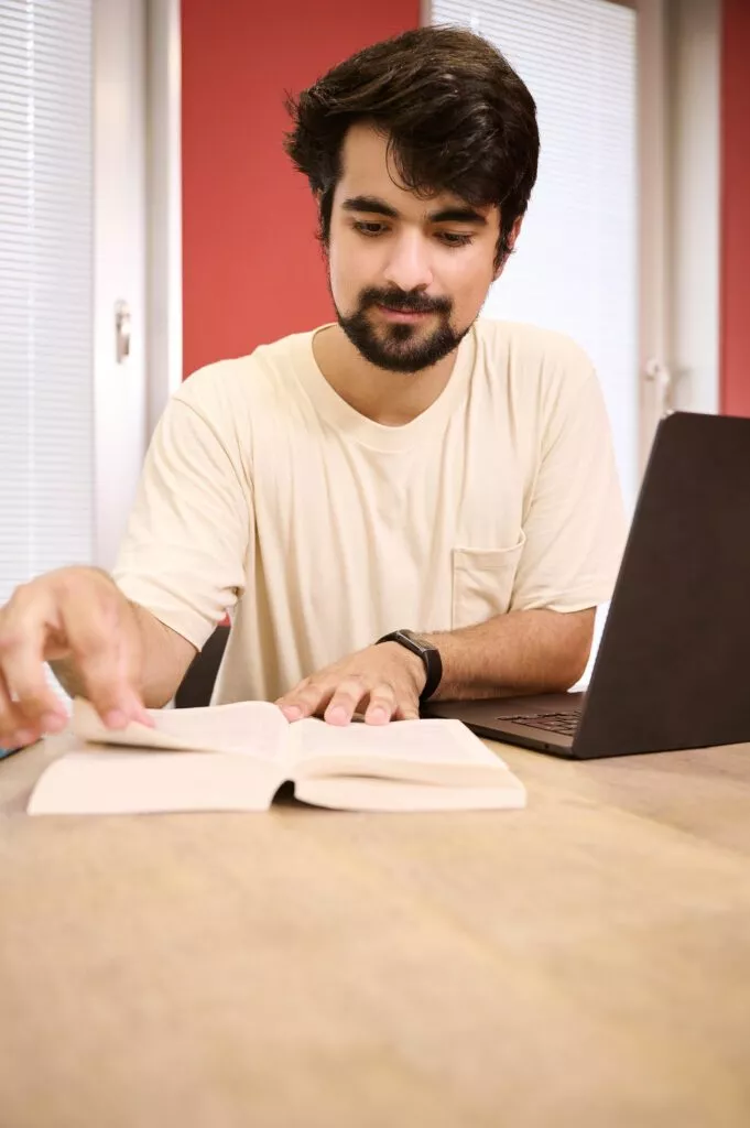 Student liest in Buch, sitzt vor dem Laptop.